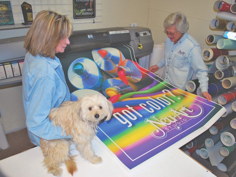 Nancy Tate, left, office manager; Dawn Neuenschwander, owner of Neu-Art Studio; and mascot Sandi check out a proof print off the wide format printer for any needed color corrections.