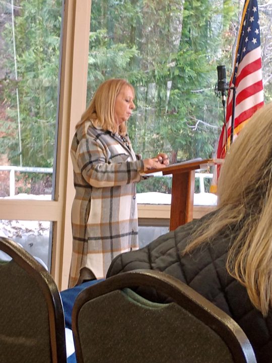 Woman speaking at podium indoors with flag nearby.