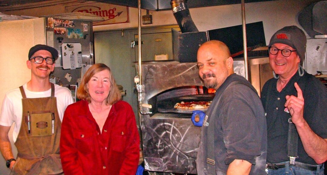 Four people in kitchen with pizza oven.