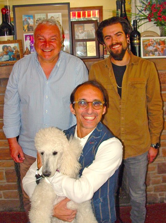 Three men smiling with a white poodle indoors.
