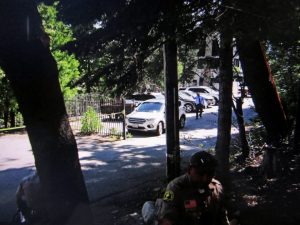 A plain-clothed investigator from the Sheriff’s Specialized Investigation Detail is seen signaling to two deputies behind trees on Land’s Edge Road on a hillside across from Lake Arrowhead Village.