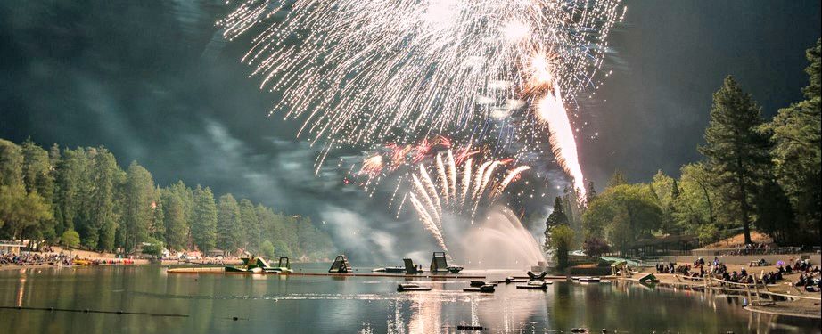 Fireworks display over forested lake with boats and people.