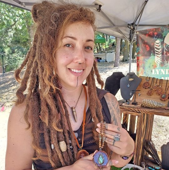Smiling artisan displaying handcrafted jewelry at market stall.