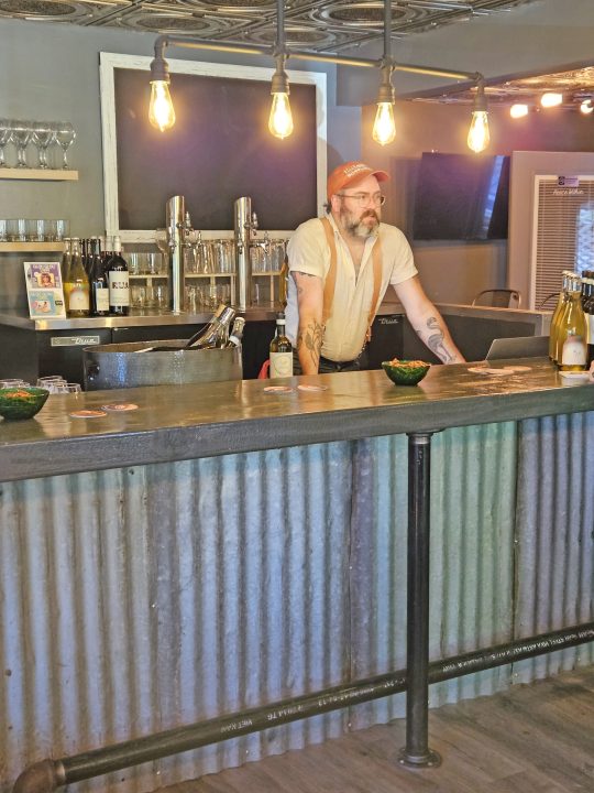 Bearded bartender standing behind rustic bar counter.