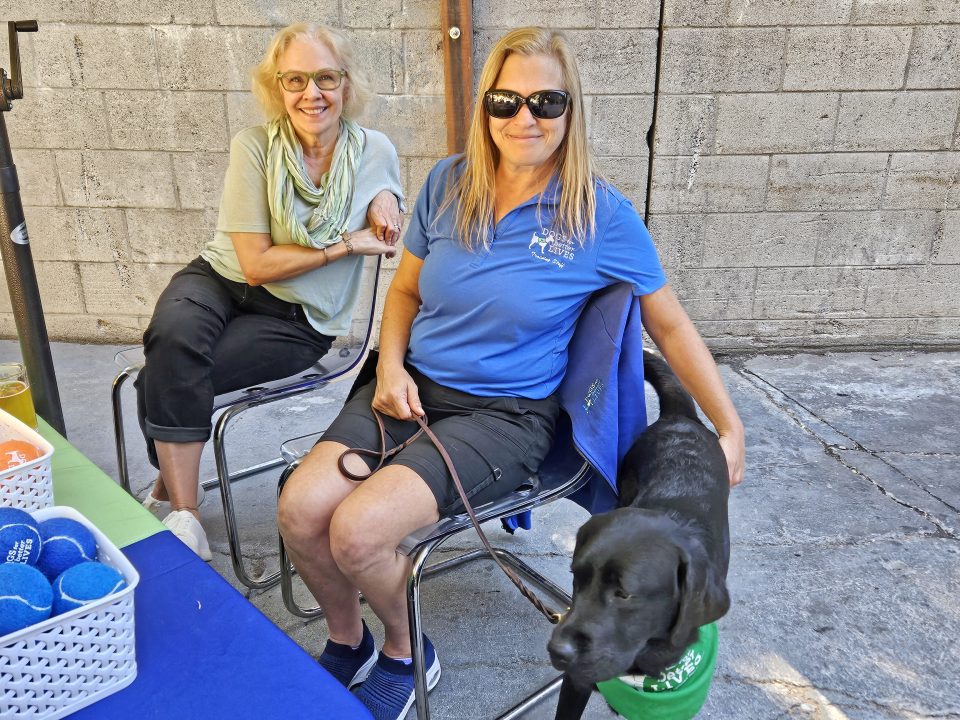 Two women with a black dog at a table.
