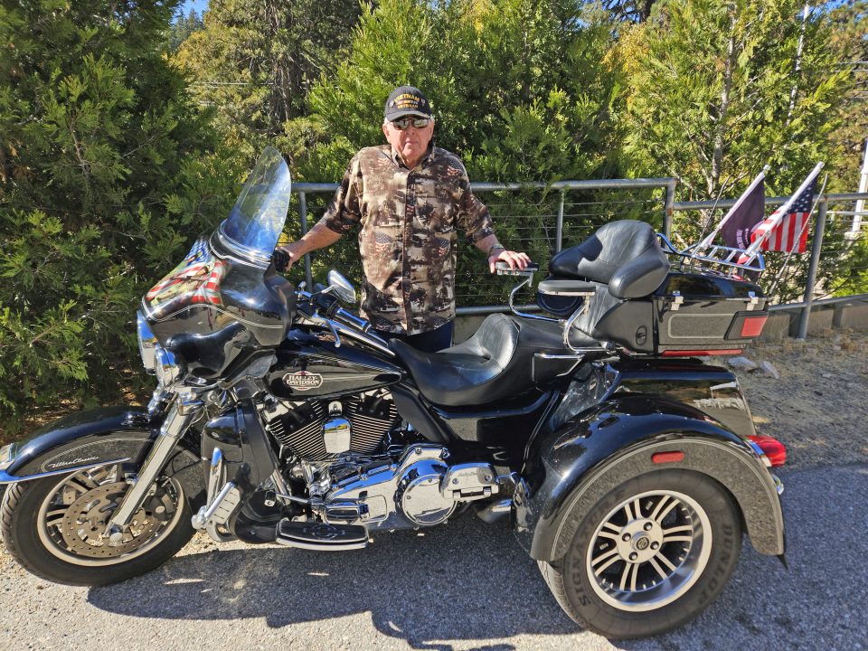 Man in camouflage beside a black motorcycle with flags.