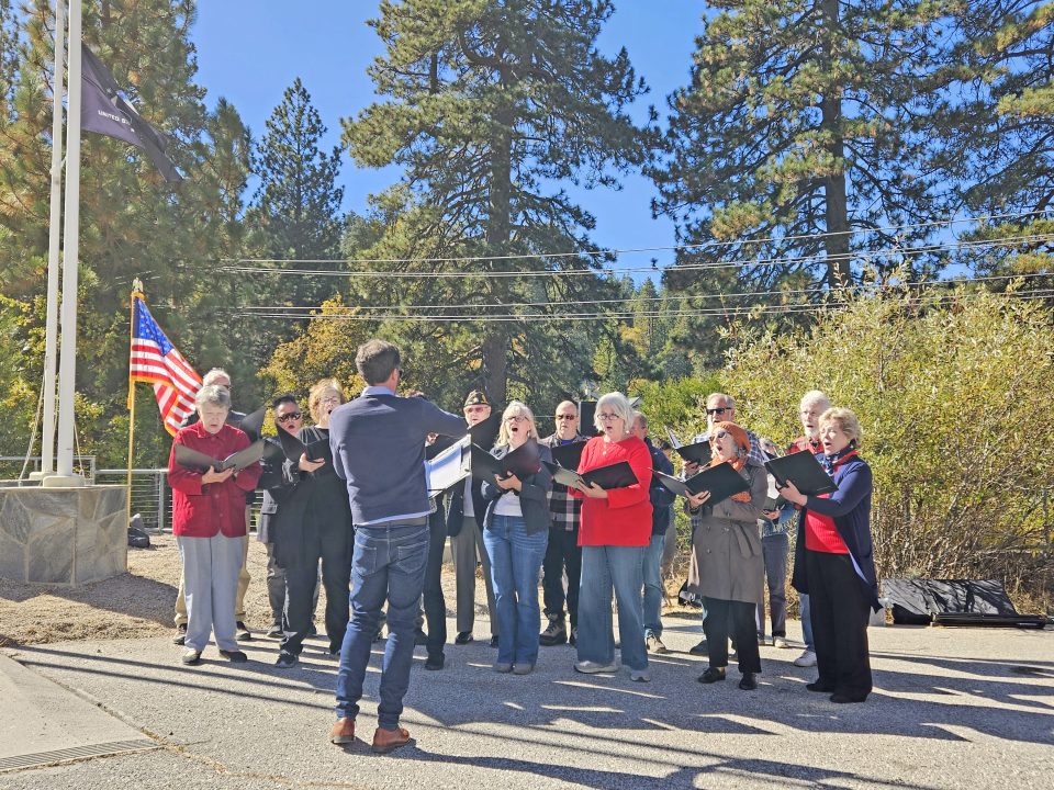 Choir performing outdoors near American flag and trees.