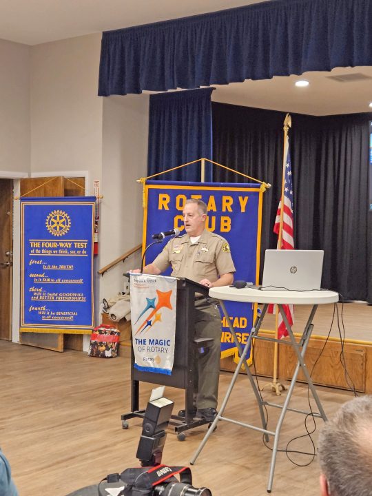 Speaker at Rotary Club meeting podium with flags.