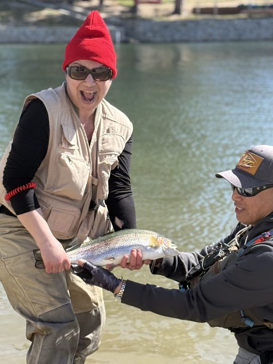 Two people holding a large fish by a lake.