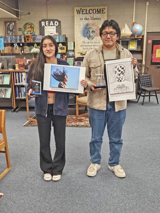 Two people holding art and trophies in library.