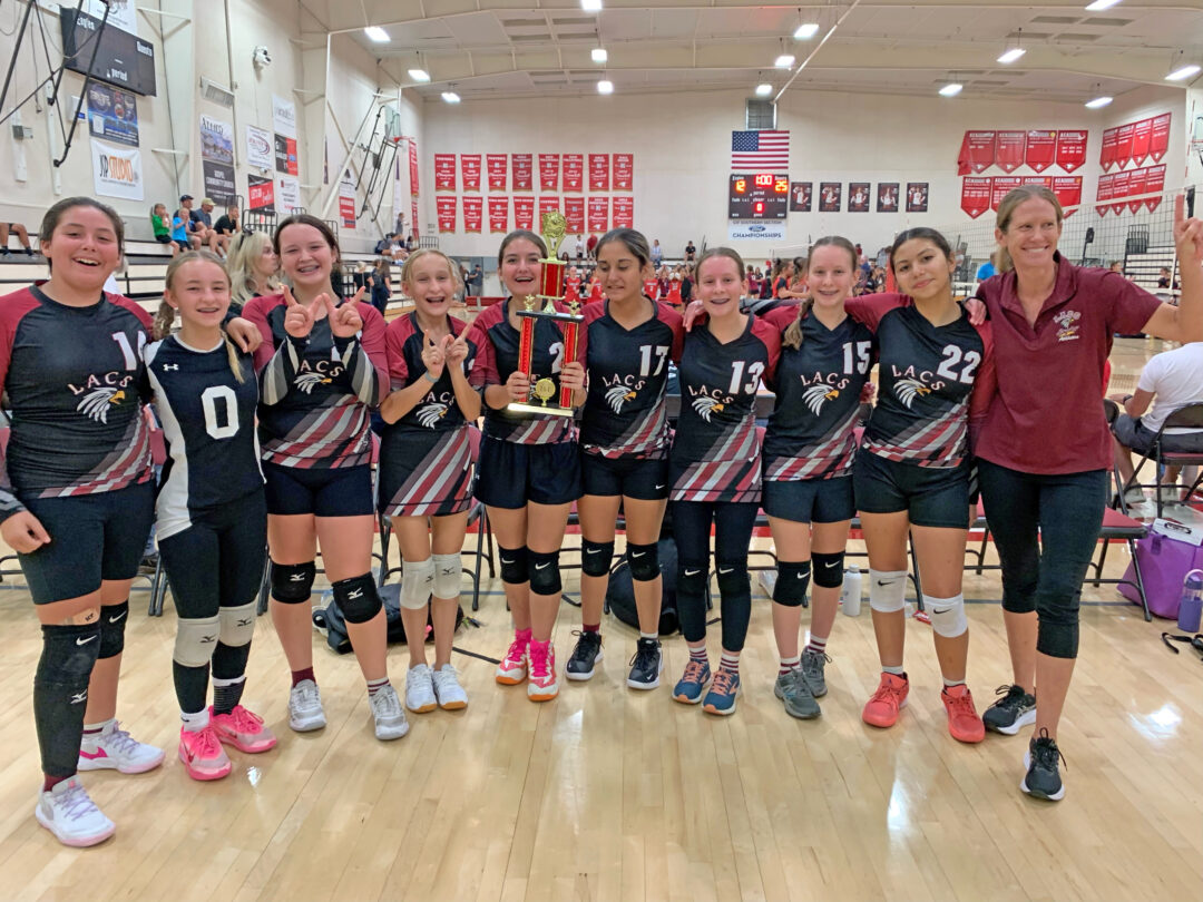 Youth volleyball team celebrates with trophy in gym.
